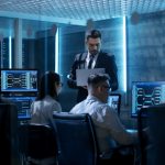 Professional IT Engineers Working in System Control Center Full of Monitors and Servers. Supervisor Holds Laptop and Holds a Briefing. Possibly Government Agency Conducts Investigation.