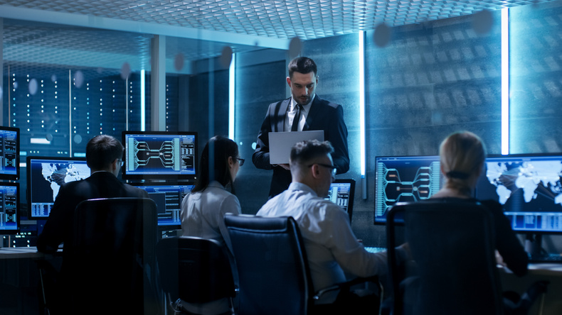 Professional IT Engineers Working in System Control Center Full of Monitors and Servers. Supervisor Holds Laptop and Holds a Briefing. Possibly Government Agency Conducts Investigation.